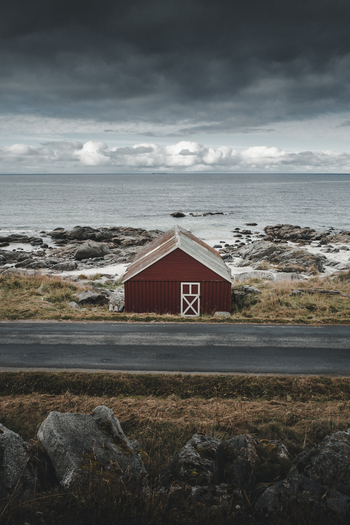 View of a red wooden cabin situated by the rocky shoreline of Vikten Beach under a dramatic cloudy sky in Vikten Nordland Norway.