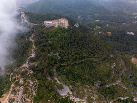 Aerial view of the Sanctuary of Sant Salvador atop a lush green mountain with a winding serpentine road and misty clouds Felanitx Illes Balears Spain. Aerial view of the Sanctuary of Sant Salvador atop a lush green mountain with a winding serpentine road and misty clouds Felanitx Illes Balears Spain.