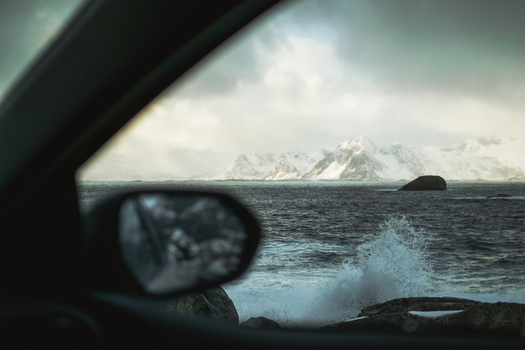 View of the rugged snow dusted mountains beyond the turbulent slate gray sea framed by the car s interior and mirror reflecting the wild scene Henningsv&aelig;r Norway.
