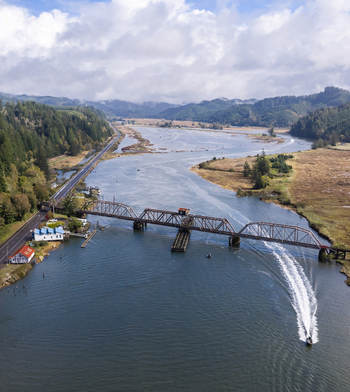 Aerial view of the Siuslaw River Train Bridge spanning the river with a boat leaving a wake surrounded by lush hills and coastal landscape in Florence Oregon United States.