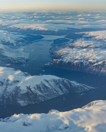 Aerial view of vast snow covered mountains and deep blue fjords under a soft golden light in Greenland. Aerial view of vast snow covered mountains and deep blue fjords under a soft golden light in Greenland.