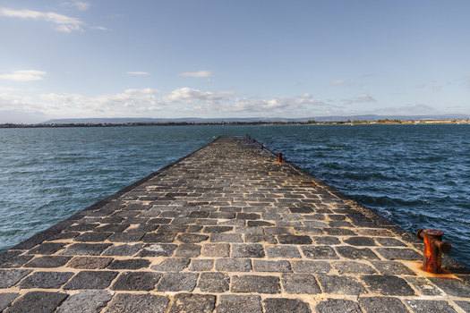 View of a cobblestone pier stretching into the turquoise sea under a clear sky rusty mooring post on the side Ortigia Sicily Italy.