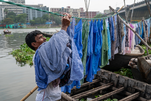 Dhaka Division Bangladesh 14 May 2026 View of a man hanging laundry on a rope over a wooden boat on the Buriganga River with urban buildings in the background.