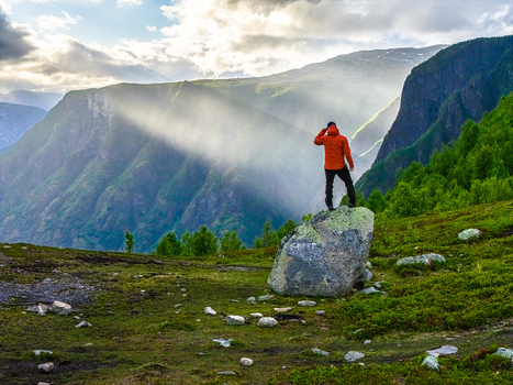 Aerial view of a person in an orange jacket standing on a large boulder overlooking the dramatic mountains and lush green valleys of the Aurlandsfjord region near Flam Vestland Norway. Aerial view of a person in an orange jacket standing on a large boulder overlooking the dramatic mountains and lush green valleys of the Aurlandsfjord region near Flam Vestland Norway.