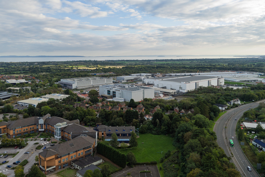 Aerial view of a large data center complex surrounded by lush greenery office buildings and a road with a distant sea view in the United Kingdom.