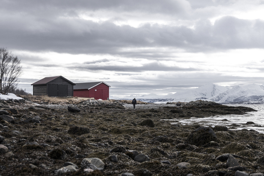 View of a person walking along a rocky shoreline with traditional wooden cabins and snow capped mountains under a cloudy sky in Tromso Troms Norway.