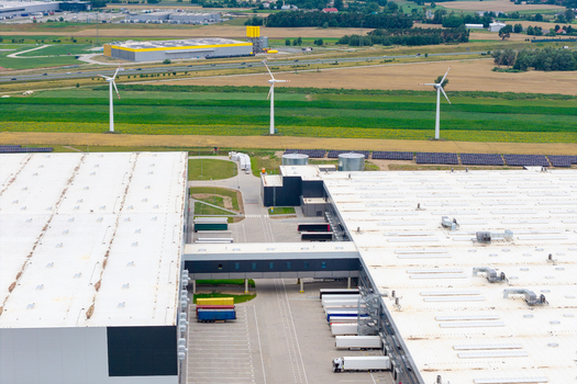 Aerial view of a large logistics distribution center with white roofs loading docks parked semi trucks and a connecting bridge near wind turbines and green fields in Poland.