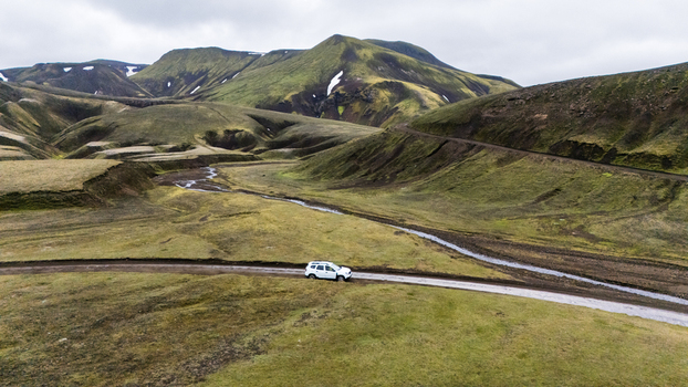 Aerial view of a white SUV driving on a dirt road through the vast green mountainous landscape of Iceland. Aerial view of a white SUV driving on a dirt road through the vast green mountainous landscape of Iceland.