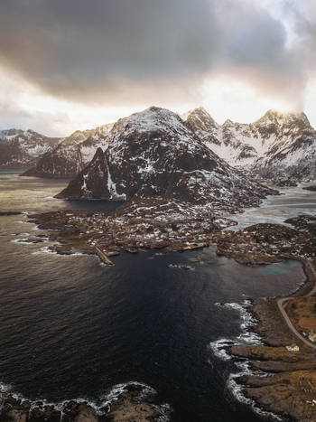 Aerial view of a quaint coastal village nestled beneath snow dusted peaks where the dark sea meets the rugged shoreline under a dramatic sky Tind Nordland Norway.