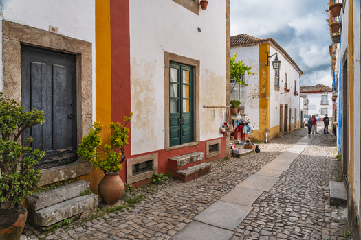 Obidos Leiria District Portugal 13 May 2026 View of historic cobblestone streets lined with traditional white houses featuring vibrant red and yellow trim green doors and potted plants. Obidos Leiria District Portugal 13 May 2026 View of historic cobblestone streets lined with traditional white houses featuring vibrant red and yellow trim green doors and potted plants.