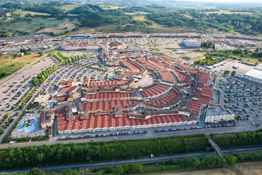 Aerial view of the Designer Outlet Serravalle featuring red roofed buildings and large parking lots surrounded by green hills in Serravalle Scrivia Piemonte Italy. Aerial view of the Designer Outlet Serravalle featuring red roofed buildings and large parking lots surrounded by green hills in Serravalle Scrivia Piemonte Italy.