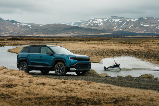 View of a blue SUV parked on a dirt track near a stream with a person kiteboarding in the water set against a backdrop of snow capped mountains in Borgarbyggd Iceland. View of a blue SUV parked on a dirt track near a stream with a person kiteboarding in the water set against a backdrop of snow capped mountains in Borgarbyggd Iceland.
