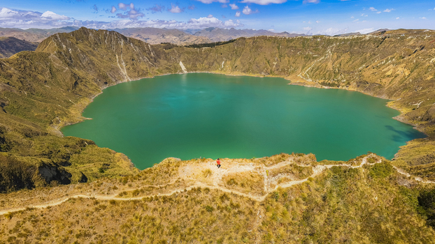 Aerial view of Quilotoa Lake with its turquoise volcanic crater water surrounded by dry grassy slopes and a person standing on the ridge in Quilotoa Cotopaxi Ecuador. Aerial view of Quilotoa Lake with its turquoise volcanic crater water surrounded by dry grassy slopes and a person standing on the ridge in Quilotoa Cotopaxi Ecuador.