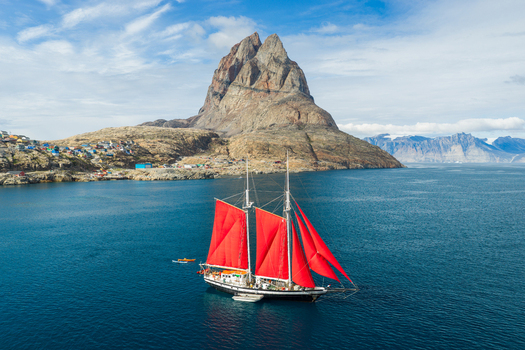 Aerial view of a sailboat with striking red sails navigating the blue waters near the iconic Uummannaq mountain and the colorful coastal town of Uummannaq Kitaa Greenland. Aerial view of a sailboat with striking red sails navigating the blue waters near the iconic Uummannaq mountain and the colorful coastal town of Uummannaq Kitaa Greenland.
