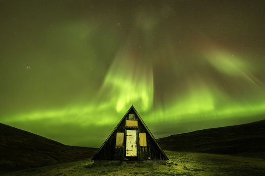 View of a small wooden A frame hut standing under a vibrant green Aurora Borealis dancing across a starry night sky over the hills of Olafsvik Snaefellsbaer Iceland.