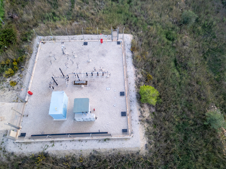 Aerial view of a fenced utility facility with industrial equipment pipes and control cabinets on a gravel surface surrounded by wild vegetation in Teulada Valencian Community Spain.