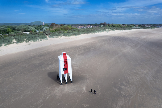 Aerial view of the historic Burnham on Sea Low Lighthouse on stilts on the sandy beach with two people walking nearby set against dunes in Burnham on Sea England United Kingdom.