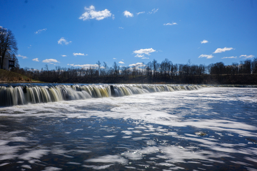 View of Ventas Rumba the widest waterfall in Europe with flowing water cascading over the limestone ledge under a clear blue sky in Kuldiga Kuldigas novads Latvia. View of Ventas Rumba the widest waterfall in Europe with flowing water cascading over the limestone ledge under a clear blue sky in Kuldiga Kuldigas novads Latvia.