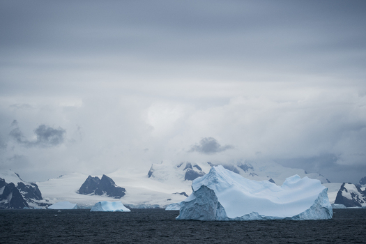 View of icebergs floating in the dark ocean with snow covered mountains of the South Orkney Islands in the background under a cloudy sky South Orkney Islands. View of icebergs floating in the dark ocean with snow covered mountains of the South Orkney Islands in the background under a cloudy sky South Orkney Islands.