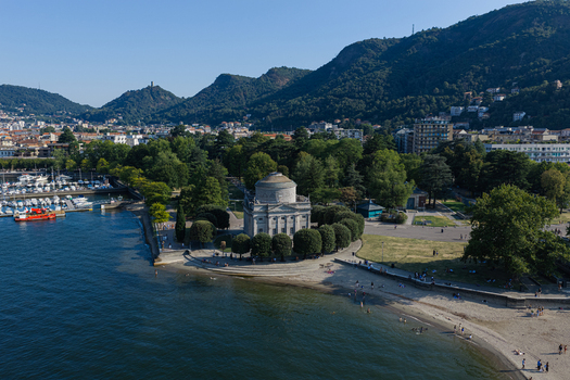 Aerial view of the Tempio Voltiano neoclassical building on the shore of Lake Como surrounded by trees and a sandy beach with people with the city and mountains in Como Lombardy Italy.