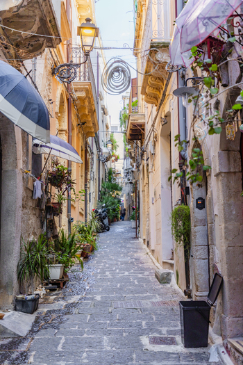 View of a quaint cobblestone alleyway lined with aged buildings potted plants and decorative umbrellas casting shadows in Ortigia Sicily Italy.