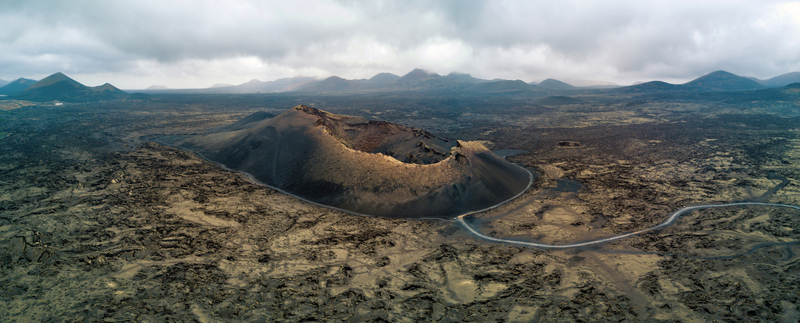 Aerial view of a massive volcanic crater surrounded by barren lava fields and a winding road under a cloudy sky Lanzarote Canarias Spain.