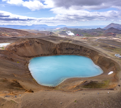 Aerial view of Krafla lake with its vibrant blue water set within a dramatic volcanic crater surrounded by rugged brown terrain and distant mountains in Thingeyjarsveit Iceland.