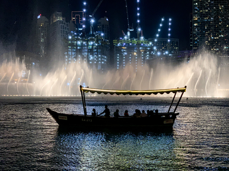 View of the Dubai Fountain at night with a traditional abra boat carrying people against a backdrop of illuminated skyscrapers and construction cranes in Dubai Dubai United Arab Emirates. View of the Dubai Fountain at night with a traditional abra boat carrying people against a backdrop of illuminated skyscrapers and construction cranes in Dubai Dubai United Arab Emirates.