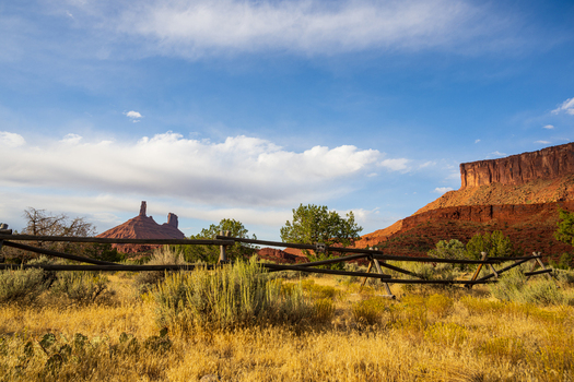View of the red rock formations along Castle Valley Drive with a rustic wooden fence in the foreground and desert vegetation under a blue sky in Grand County Utah United States.