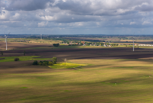 Aerial view of wind turbines standing in vast agricultural fields with patches of green and brown earth under a cloudy sky in Pomeranian Voivodeship Poland.