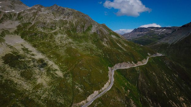 Aerial view of the winding Furka Pass road carved into steep green mountain slopes under a blue sky in Obergoms Valais Switzerland. Aerial view of the winding Furka Pass road carved into steep green mountain slopes under a blue sky in Obergoms Valais Switzerland.