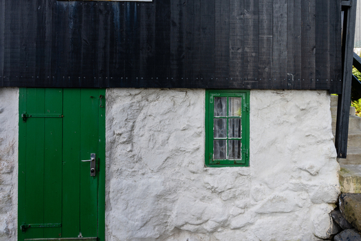 View of a traditional house with white stone walls black wooden siding a green door and a green framed window in the Faroe Islands. View of a traditional house with white stone walls black wooden siding a green door and a green framed window in the Faroe Islands.