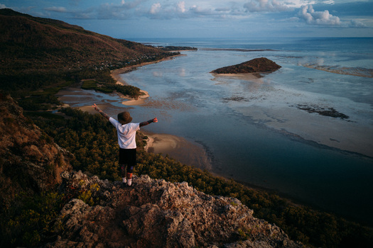 Aerial view of a person standing on a rocky cliff overlooking the turquoise lagoon and lush mountains during sunset in Moka District Mauritius. Aerial view of a person standing on a rocky cliff overlooking the turquoise lagoon and lush mountains during sunset in Moka District Mauritius.