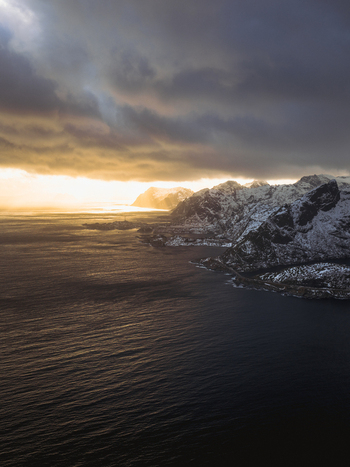Aerial view of the rugged coastline meets the dark rippling sea under a sky streaked with gold and ominous clouds Reinebringen Nordland Norway.