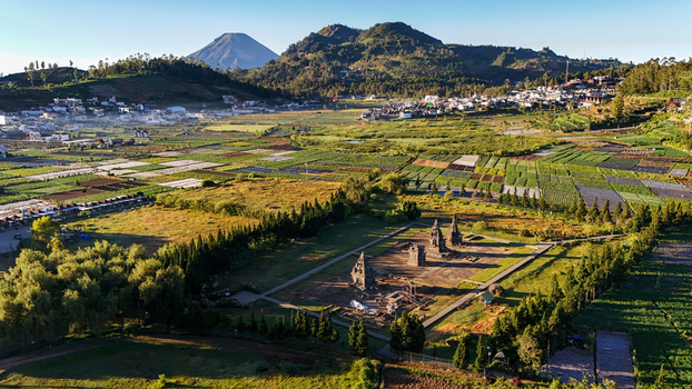 Aerial view of Candi Arjuna temple complex surrounded by green agricultural fields and a village with volcanic mountains in the background Kecamatan Batur Jawa Tengah Indonesia.