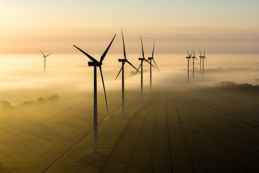 Aerial view of wind turbines standing tall against a backdrop of thick fog casting long shadows across the golden fields Wislina Pomeranian Voivodeship Poland. Aerial view of wind turbines standing tall against a backdrop of thick fog casting long shadows across the golden fields Wislina Pomeranian Voivodeship Poland.