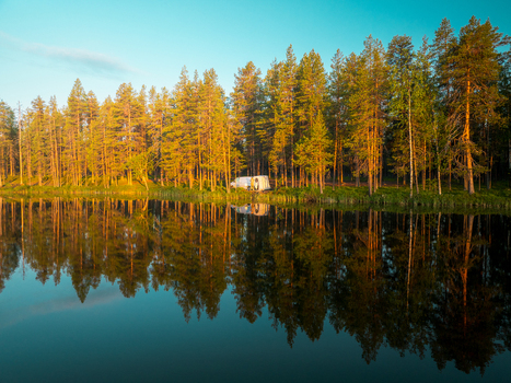 Aerial view of a campervan parked near a dense pine forest on the edge of a calm lake in northern Sweden with golden hour sunlight reflecting on the water surface. Aerial view of a campervan parked near a dense pine forest on the edge of a calm lake in northern Sweden with golden hour sunlight reflecting on the water surface.
