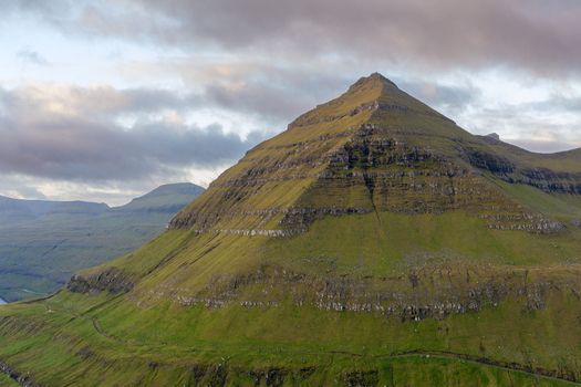 View of a pyramid shaped mountain with lush green slopes and rocky strata under a cloudy sky in the Faroe Islands. View of a pyramid shaped mountain with lush green slopes and rocky strata under a cloudy sky in the Faroe Islands.