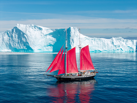 Aerial view of a sailboat with vibrant red sails navigating through deep blue waters in front of a massive towering iceberg Greenland. Aerial view of a sailboat with vibrant red sails navigating through deep blue waters in front of a massive towering iceberg Greenland.