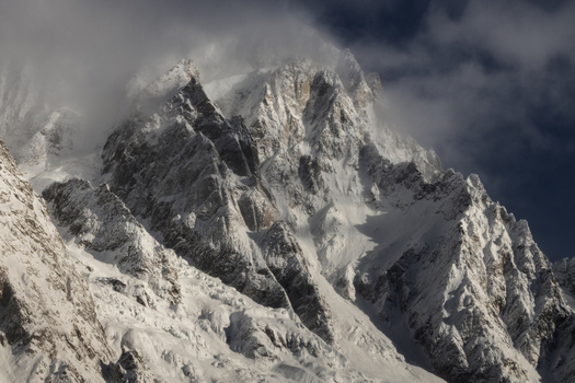 View of sharp snow covered mountain peaks and rocky ridges partially hidden by thick clouds under a dark sky in Aosta Valley Italy. View of sharp snow covered mountain peaks and rocky ridges partially hidden by thick clouds under a dark sky in Aosta Valley Italy.