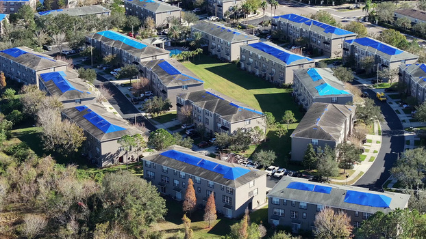 Aerial view of a residential apartment complex with blue tarps covering damaged roofs surrounded by green lawns trees and parking areas in Tampa Florida United States.