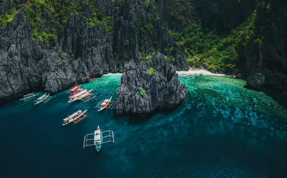 Aerial view of limestone cliffs surrounding a turquoise lagoon with traditional outrigger boats floating in the clear water in El Nido Mimaropa Philippines. Aerial view of limestone cliffs surrounding a turquoise lagoon with traditional outrigger boats floating in the clear water in El Nido Mimaropa Philippines.