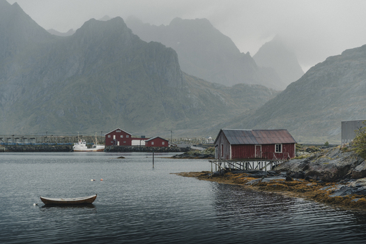 Sund Nordland Norway 04 September 2025 View of red wooden rorbuer cabins on stilts along the rocky shoreline with a small boat in the calm water against misty mountains.