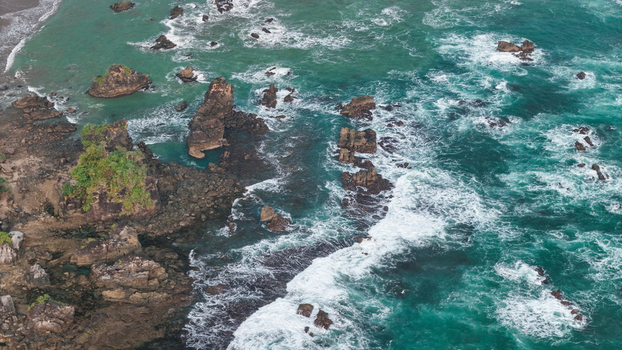 Aerial view of a rugged coastline with dark volcanic rocks turquoise ocean waves crashing against the shore and lush green vegetation on the cliffs in Indonesia. Aerial view of a rugged coastline with dark volcanic rocks turquoise ocean waves crashing against the shore and lush green vegetation on the cliffs in Indonesia.