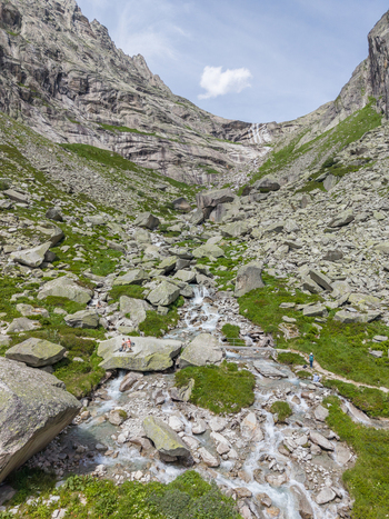 Aerial view of a mountain creek flowing through rocky terrain with a small footbridge a hiking trail and people resting on a large boulder in Switzerland.