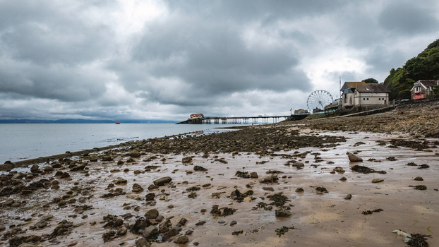 View of Mumbles Pier stretching into the sea with a Ferris wheel and rocky beach under a cloudy sky Mumbles Wales United Kingdom.