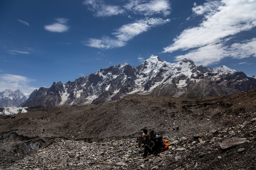 View of people resting on the rocky moraine of the Baltoro Glacier with snow capped mountain peaks under a blue sky in Baltoro Gilgit Baltistan Pakistan.