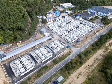 Aerial view of a large industrial battery energy storage system with rows of white containers adjacent to a dense forest and a road in Poland.