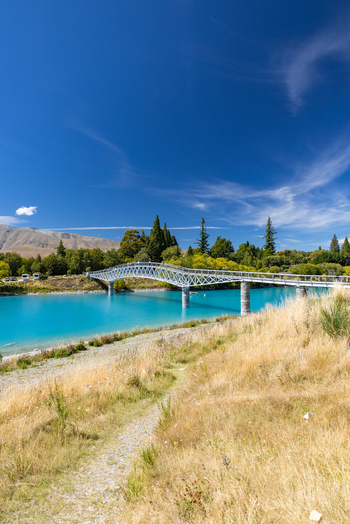 View of a brilliant turquoise river flowing beneath a sleek modern bridge contrasting with the golden grasses under a clear blue sky Lake Tekapo New Zealand. View of a brilliant turquoise river flowing beneath a sleek modern bridge contrasting with the golden grasses under a clear blue sky Lake Tekapo New Zealand.