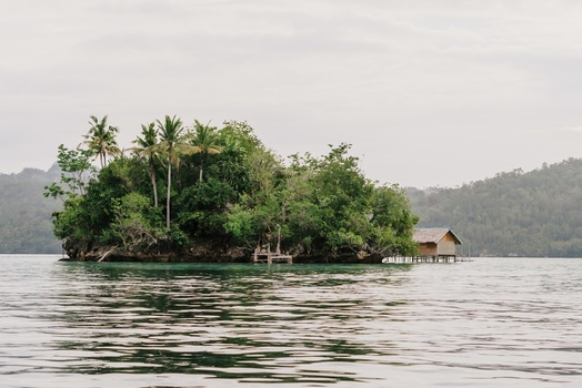 View of Raja Ampat featuring a small tropical island with lush vegetation palm trees and a wooden stilt house over calm water in Southwest Papua Indonesia. View of Raja Ampat featuring a small tropical island with lush vegetation palm trees and a wooden stilt house over calm water in Southwest Papua Indonesia.